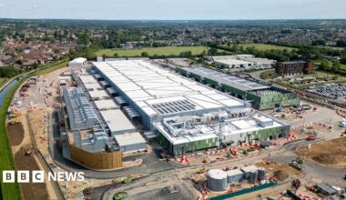 An aerial view of a data centre in Hertfordshire, it is a long white and silver square building, with a newly build road around the outside and construction still going on. To the left is a canal and int he background green fields and a settlement beyond that.