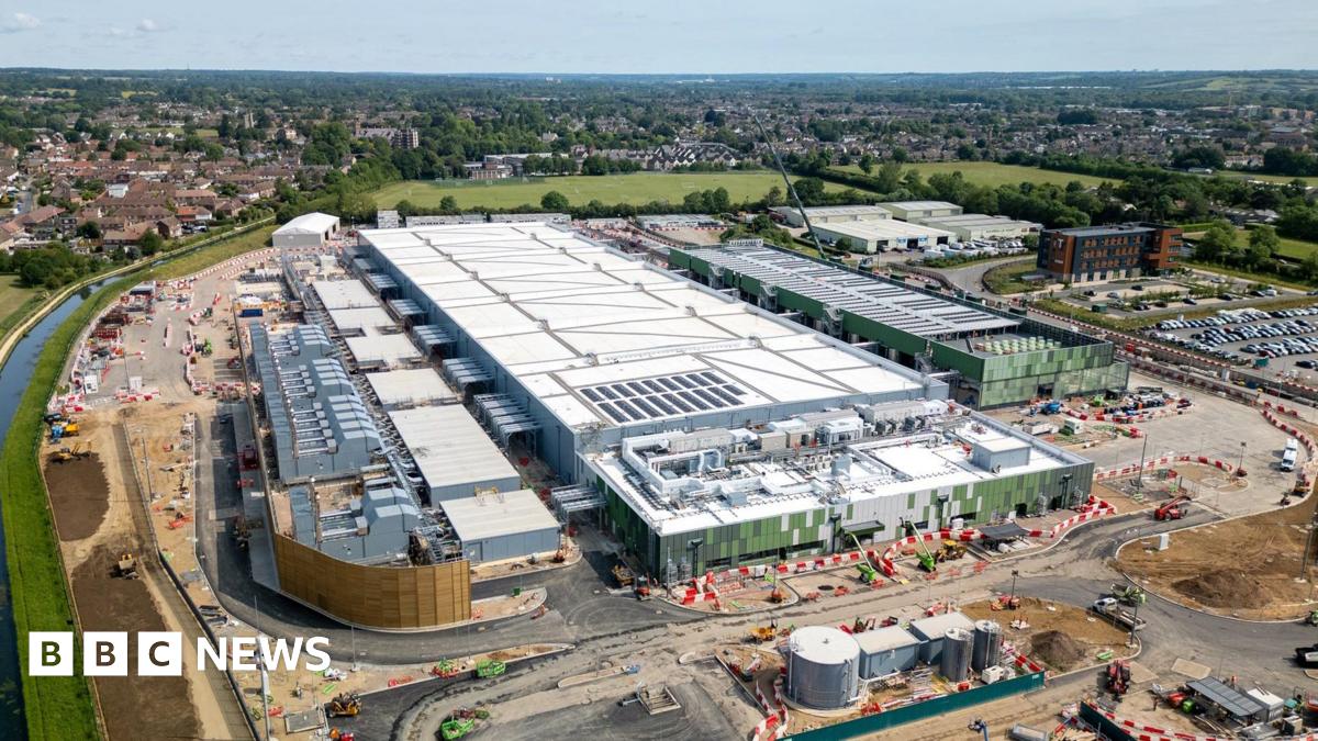 An aerial view of a data centre in Hertfordshire, it is a long white and silver square building, with a newly build road around the outside and construction still going on. To the left is a canal and int he background green fields and a settlement beyond that.