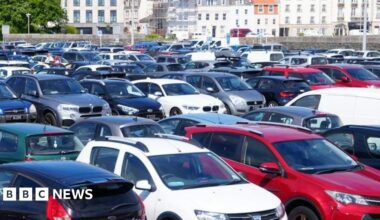 Cars fill all the spaces at a car park with a wall and harbour buildings beyond.