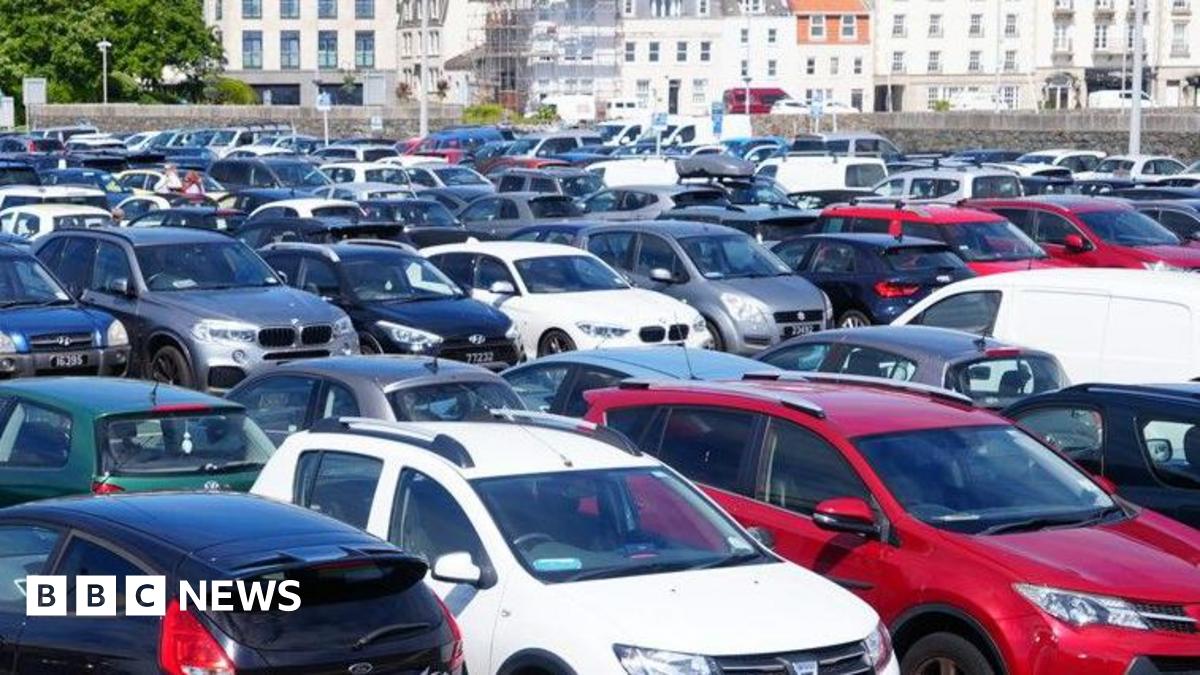 Cars fill all the spaces at a car park with a wall and harbour buildings beyond.