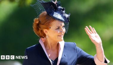 Sarah Ferguson wears a black and white checked jacket with a black top underneath, and is sitting against a black background - plants can just be seen at the side. Her red hair is pushed back with a thin black headband and she has a neutral expression on her face