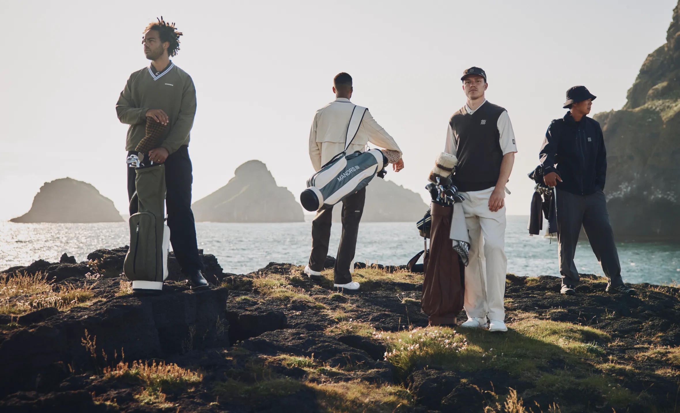Four men in golf attire and carrying golf bags stand on a rocky cliff overlooking the ocean with small islands in the distance.