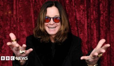A man with long brown hair and red sunglasses with his hands outstretched and a black top in front of a red curtain