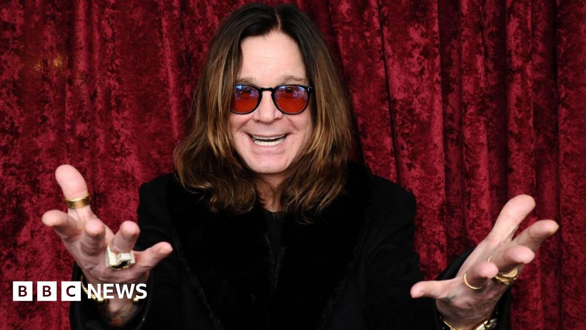 A man with long brown hair and red sunglasses with his hands outstretched and a black top in front of a red curtain
