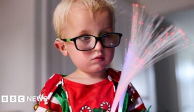 Five-year-old Harvey Hind with blond hair wearing black glasses in a red, white  and green Christmas top with candy canes holds two sensory optic wands which he is looking at.