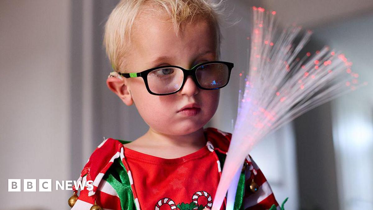 Five-year-old Harvey Hind with blond hair wearing black glasses in a red, white  and green Christmas top with candy canes holds two sensory optic wands which he is looking at.