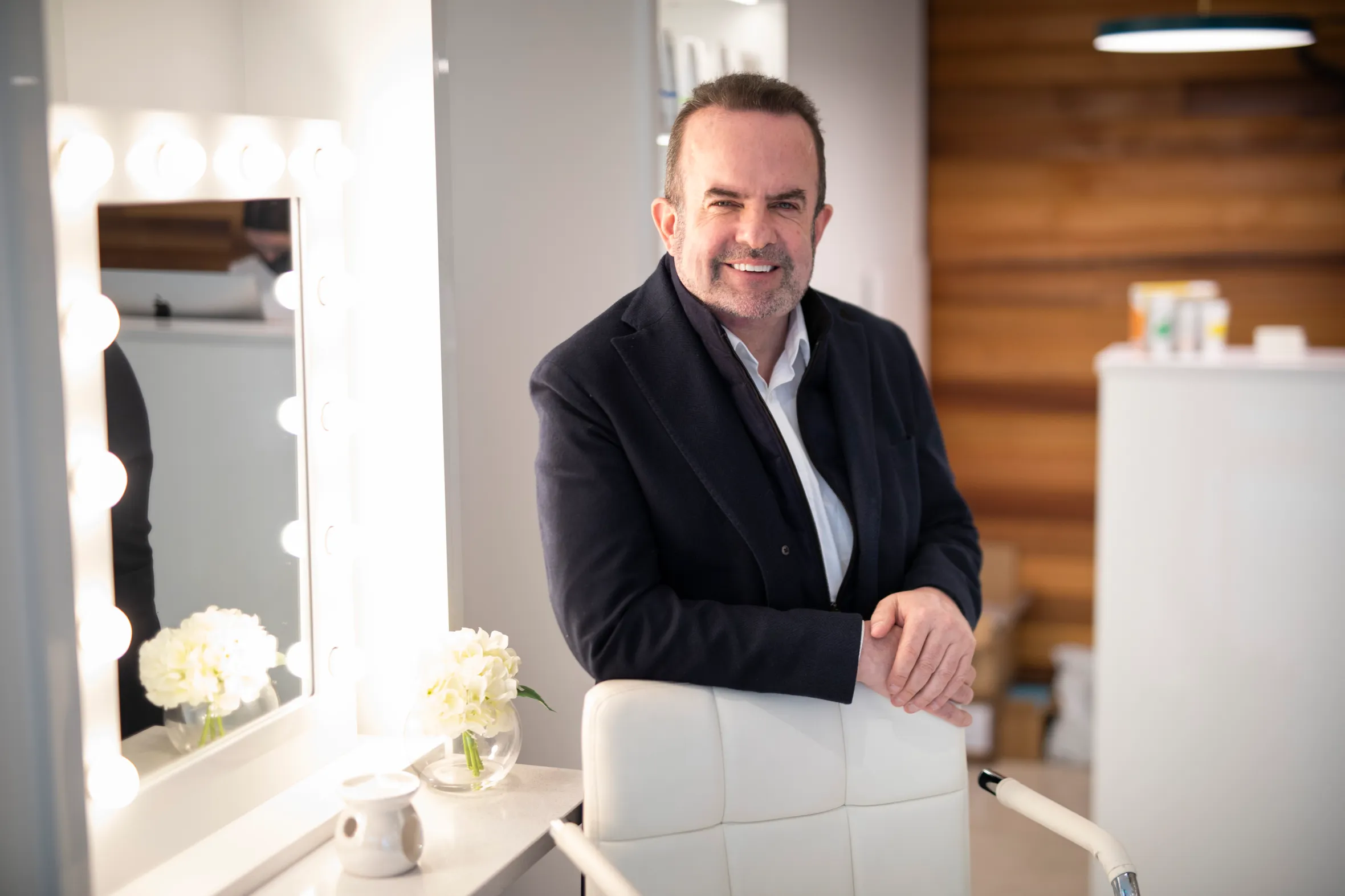 Pat Phelan, Co-Founder at SISU Aesthetic Clinic, stands next to a white chair with his hands resting on its back.