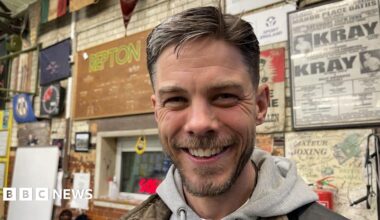 A man with brown hair and a beard stands smiling at the camera in a boxing club. He is wearing a grey hooded sweatshirt. There are posters on the wall behind him with words like Kray, amateur boxing and Repton. There is also a small booth-style window with a red digital clock in front of the glass.