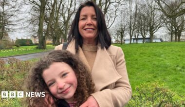 Louise James stands behind her daughter Rioghnach. Louise has long dark hair and is wearing a tan woollen coat over a tan woollen sweater. She has her arms on Rioghnach who is smiling and has curly brown hair. They are standing in a park, trees and a rolling stretch of grass can be seen behind them.