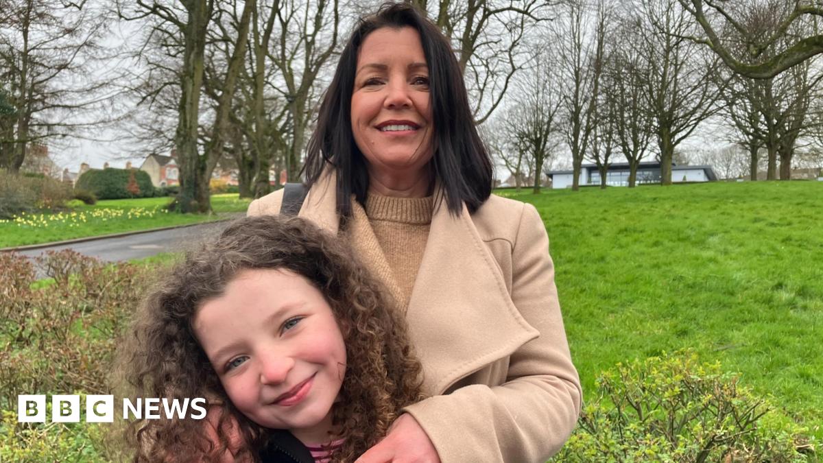 Louise James stands behind her daughter Rioghnach. Louise has long dark hair and is wearing a tan woollen coat over a tan woollen sweater. She has her arms on Rioghnach who is smiling and has curly brown hair. They are standing in a park, trees and a rolling stretch of grass can be seen behind them.