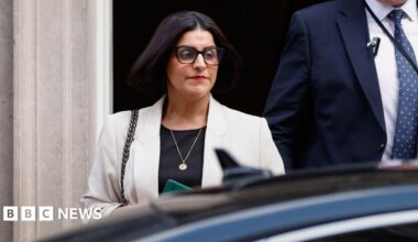 Home Secretary Shabana Mahmood walking to a car wearing dark brown glasses, red lipstick, and a light grey blazer.