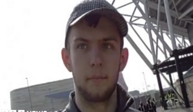 A man with short dark hair and a beard in a grey top and grey cap in front of a large grey building and the corner of a football stand
