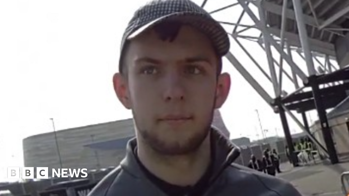 A man with short dark hair and a beard in a grey top and grey cap in front of a large grey building and the corner of a football stand