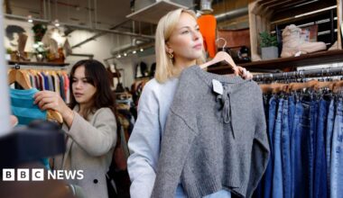 A young blonde woman in a shop holding a grey jumper up against herself