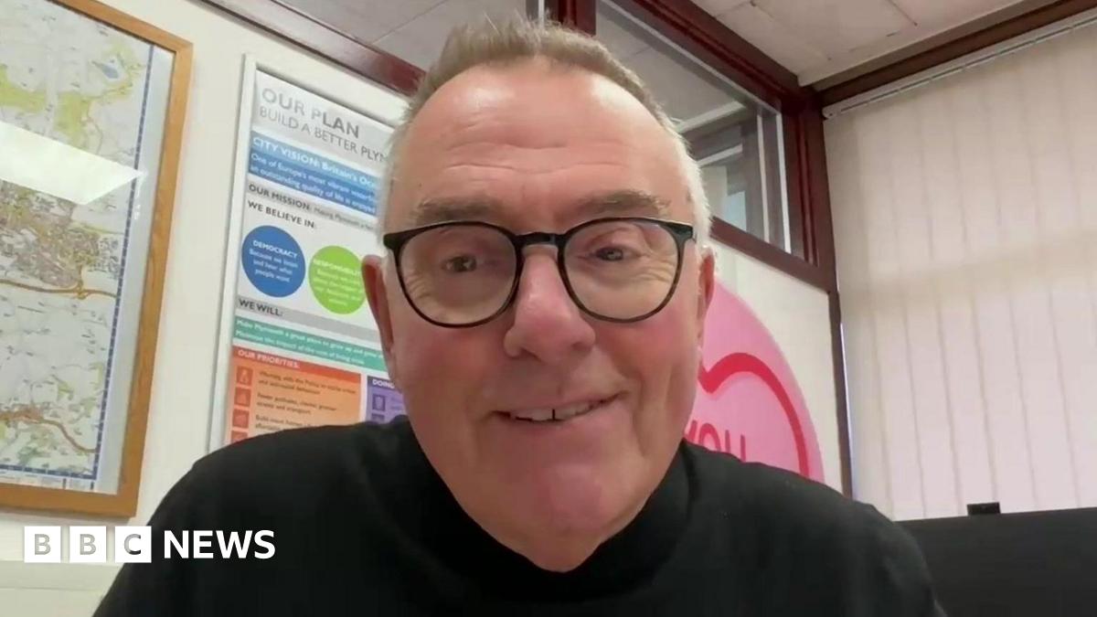 Plymouth City Council leader Tudor Evans smiles. He is wearing a black jumper and has black rimmed glasses on. He is sat in an office with posters and a map of Plymouth on the walls.