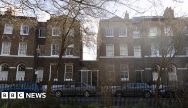 A row of terraced houses, all of which are three storeys high apart from number 25 and 26 which stand at one storey with their front doors next to each other.