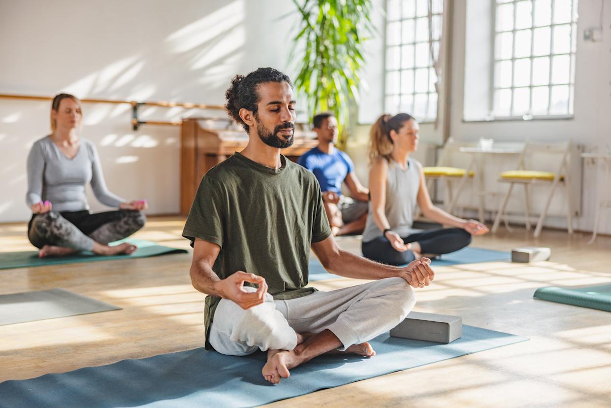 An adult Arab male with a ponytail meditating in a yoga class. He is surrounded by other yoga class participants that are diverse in age, gender and race. The yoga practitioner looks calm and relaxed. The class in taking place in a cosy bright studio with big windows and plants.