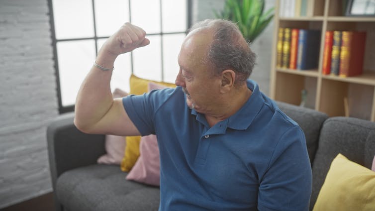An older many sitting on a couch flexes his biceps.