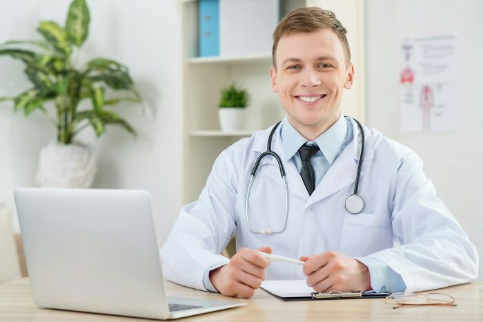 Smiling male doctor with stethoscope, sitting at desk with laptop and clipboard, discussing patient cases of faking illness.