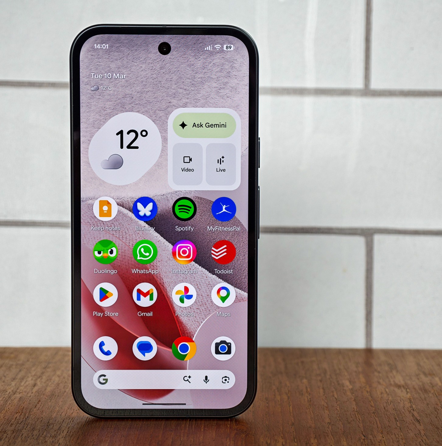 Photo of Google Pixel 10A in front of white tiles, showing the home screen