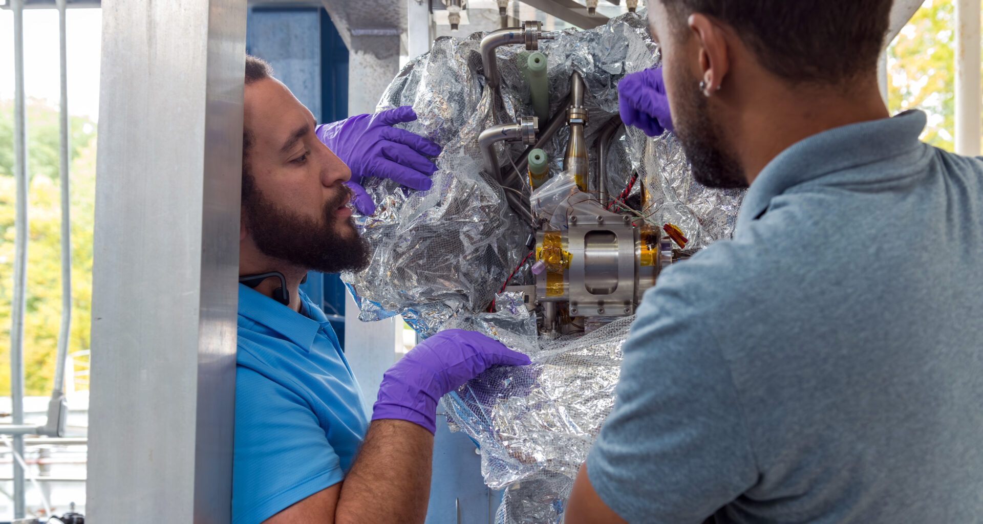Two engineers wearing purple gloves peel away wrapping around metal and yellow hardware. The hardware is mounted inside a steel metal frame. This technology could help NASA fuel future missions,