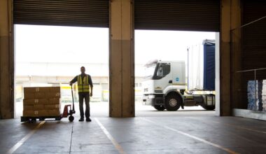 A worker wearing a yellow vest stands looking into an empty warehouse with a cart full of food boxes next to him and a truck in the background