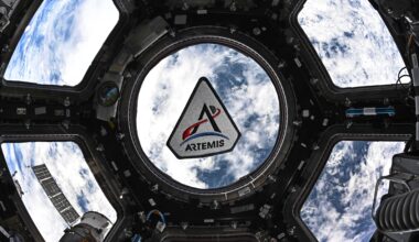 The Artemis program patch in the space station's cupola. The patch is framed by the center circular window in the space. Earth can be seen behind the patch. The Artemis patch has five sides. It has a white background with a black border. There's a black "A," for Artemis. The crossbar of the A is made by a curving red trajectory line that crosses from left to right. That line continues beyond the "A" and beyond a small gray circle representing the Moon. Below the "A" is a blue crescent representing Earth. At the very bottom is the word "Artemis."