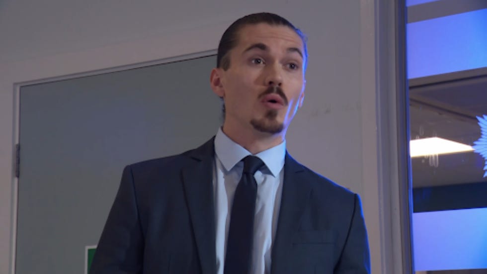 hollyoaks sully person standing in a formal suit in an indoor setting