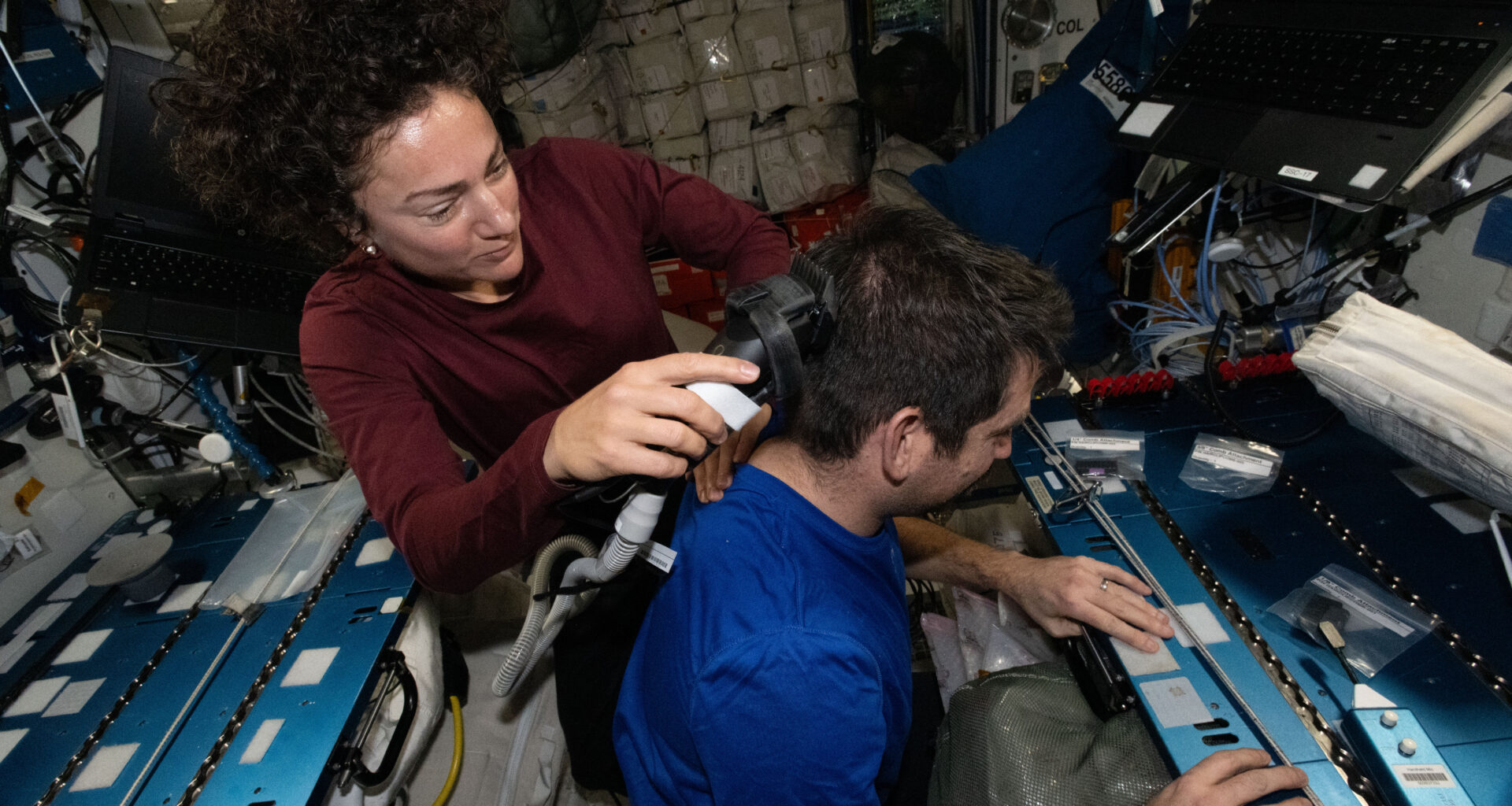 Jessica Meir holds an electric razor with a vacuum to Jack Hathaway's hair. She looks down at what she's doing, while Hathaway faces to the right of the image. Meir's brown curly hair floats up above her head.