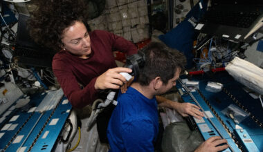 Jessica Meir holds an electric razor with a vacuum to Jack Hathaway's hair. She looks down at what she's doing, while Hathaway faces to the right of the image. Meir's brown curly hair floats up above her head.