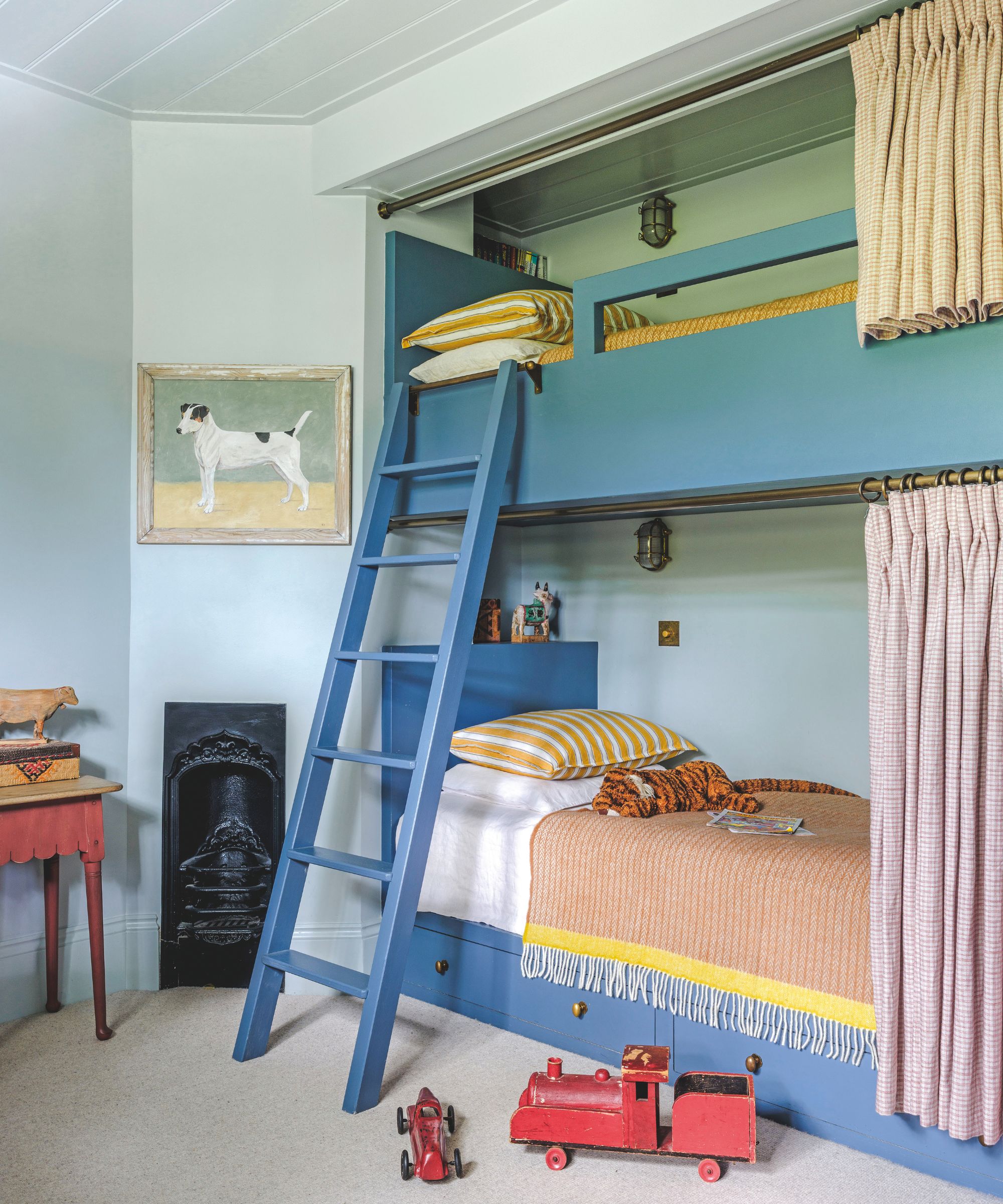 A cozy children's room with built-in blue bunk beds, an integrated wooden ladder, and yellow striped pillows set against a pale blue wall with a small fireplace.