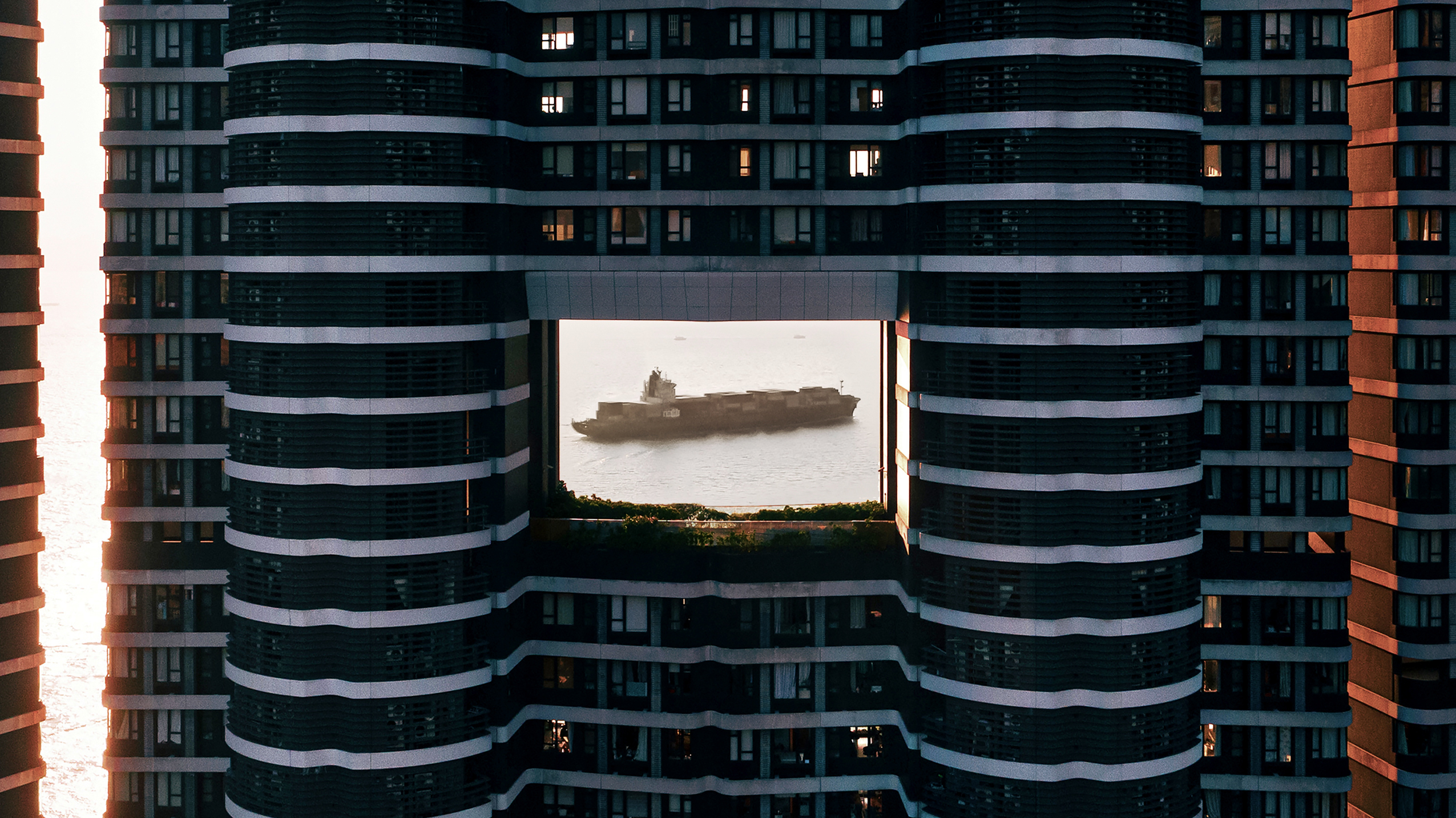 Close up of: A ship passes by in the sea, viewed through a rectangular opening between two tall, dark skyscrapers at sunset