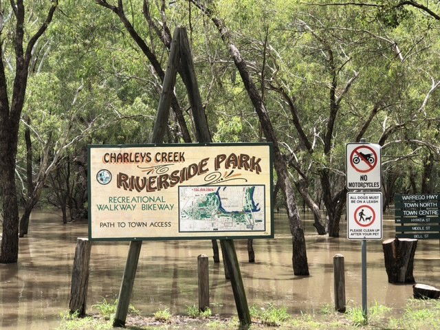 A sign reads charleys creek riverside park in front of a creek with muddy waters. 