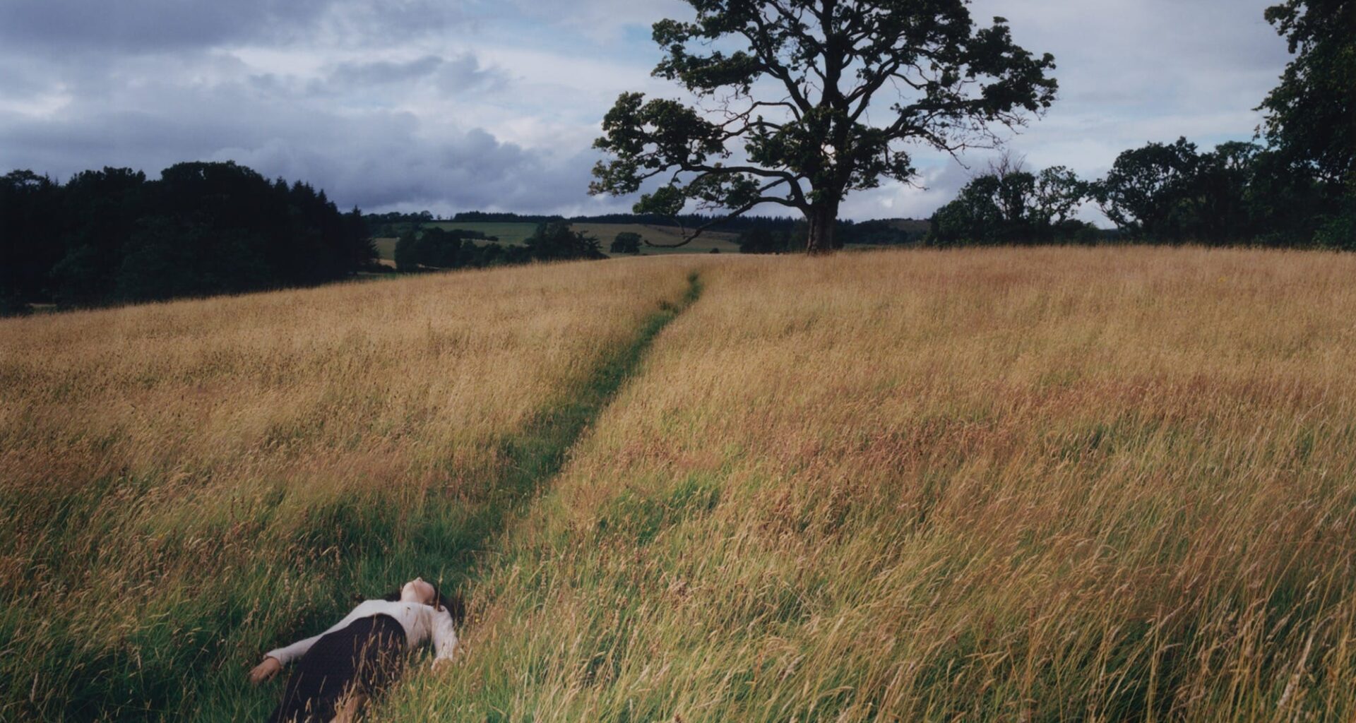 A fine art photograph by Camille Lemoine of a young woman laying in a trodden path in a meadow with a tree in the background