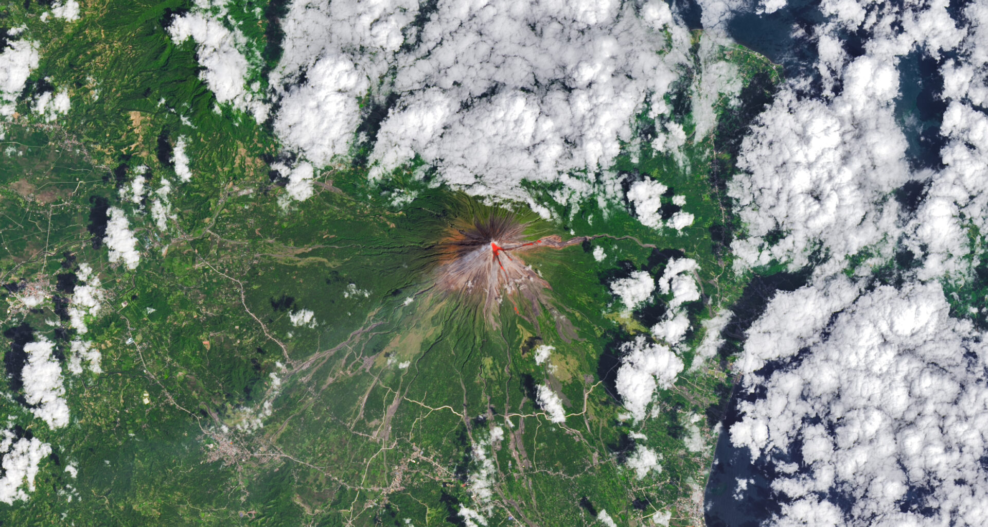 The upper slopes of Mayon volcano appear brown, with several narrow channels radiating from the crater. A red infrared heat signature appears near the summit, with red streaks extending east and southeast. The lower slopes are green and forested. Farmland and towns are visible in the lower part of the image.