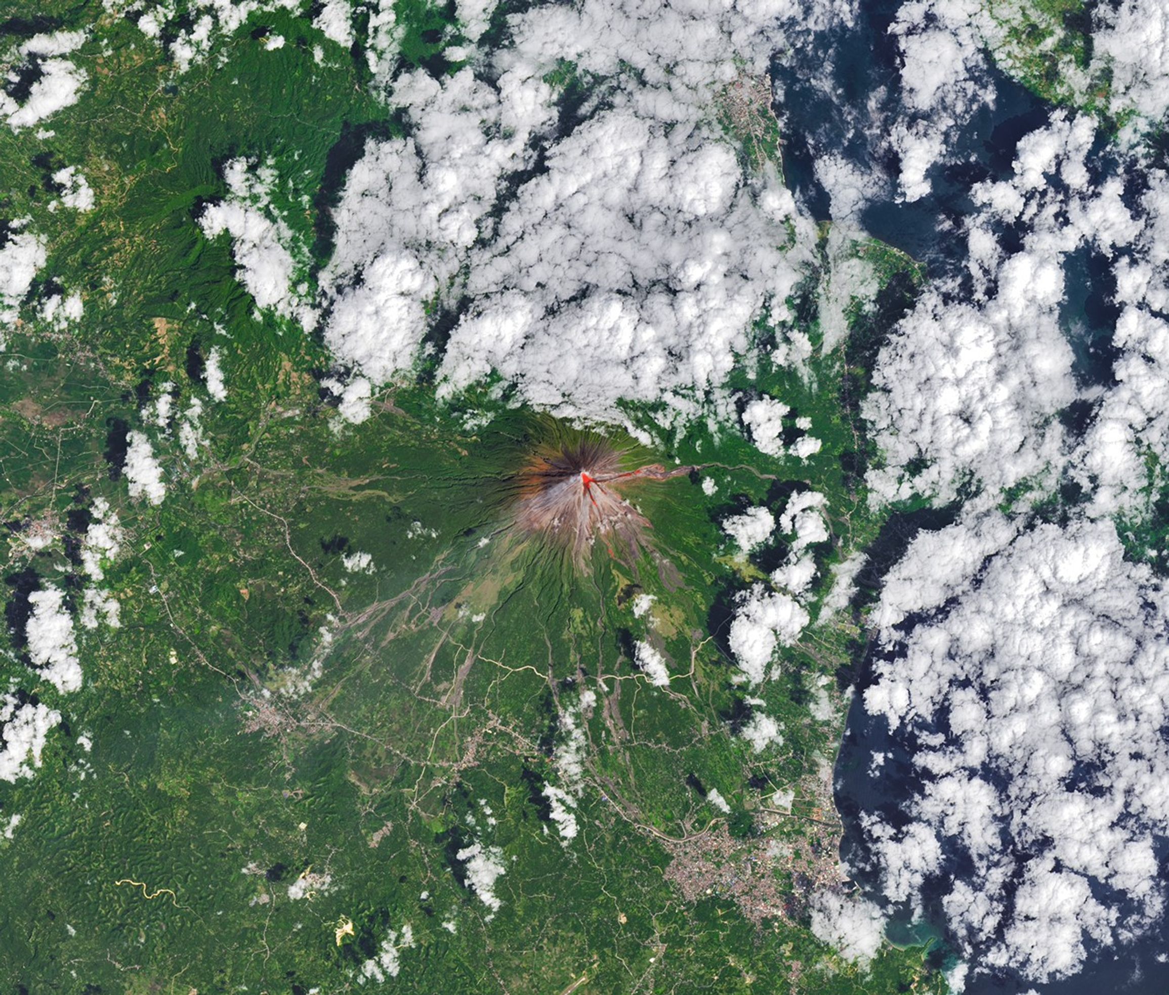 The upper slopes of Mayon volcano appear brown, with several narrow channels radiating from the crater. A red infrared heat signature appears near the summit, with red streaks extending east and southeast. The lower slopes are green and forested. Farmland and towns are visible in the lower part of the image.