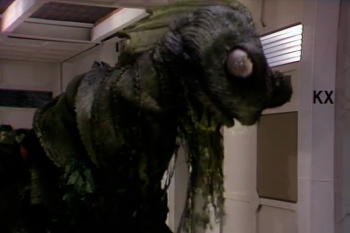 The Myrka in Doctor Who: Warriors of the Deep, a large, slimy green alien creature with ridged skin and a bulging eye looming through a corridor hatch, dripping goo and filling most of the frame.