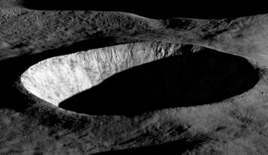 Oblique view of a large, circular lunar crater with steep, rugged walls. Sunlight illuminates one side of the crater rim and inner wall, while the crater floor and much of the interior remain in deep shadow. The surrounding terrain appears gray and heavily textured with smaller craters and rocky ridges. The black sky of space fills the background.