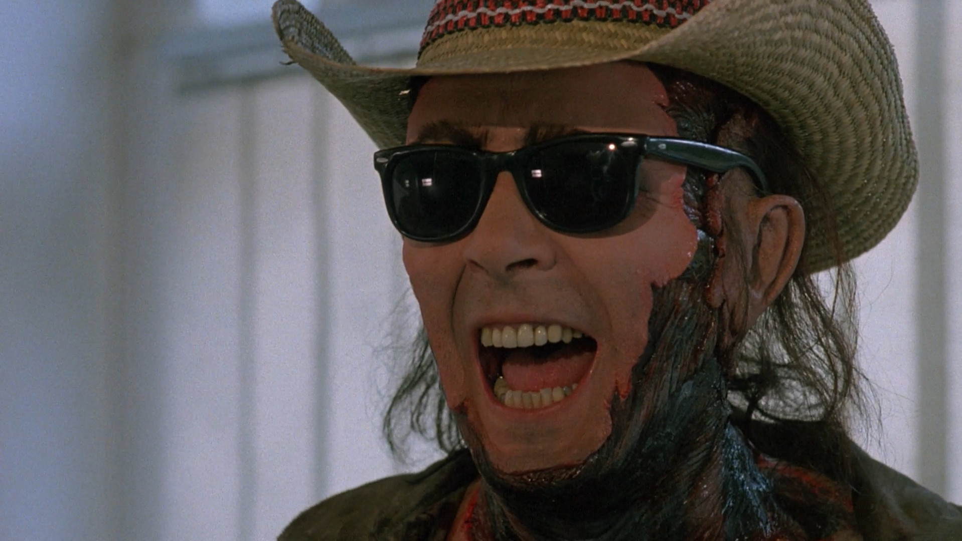 A man whose face is mostly torn off wearing sunglasses and a cowboy hat laughing.
