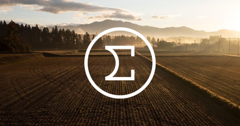 A plowed field at sunrise with mountains in the background, trees lining the horizon, and a large white Sigma symbol enclosed in a circle overlaying the center of the image.
