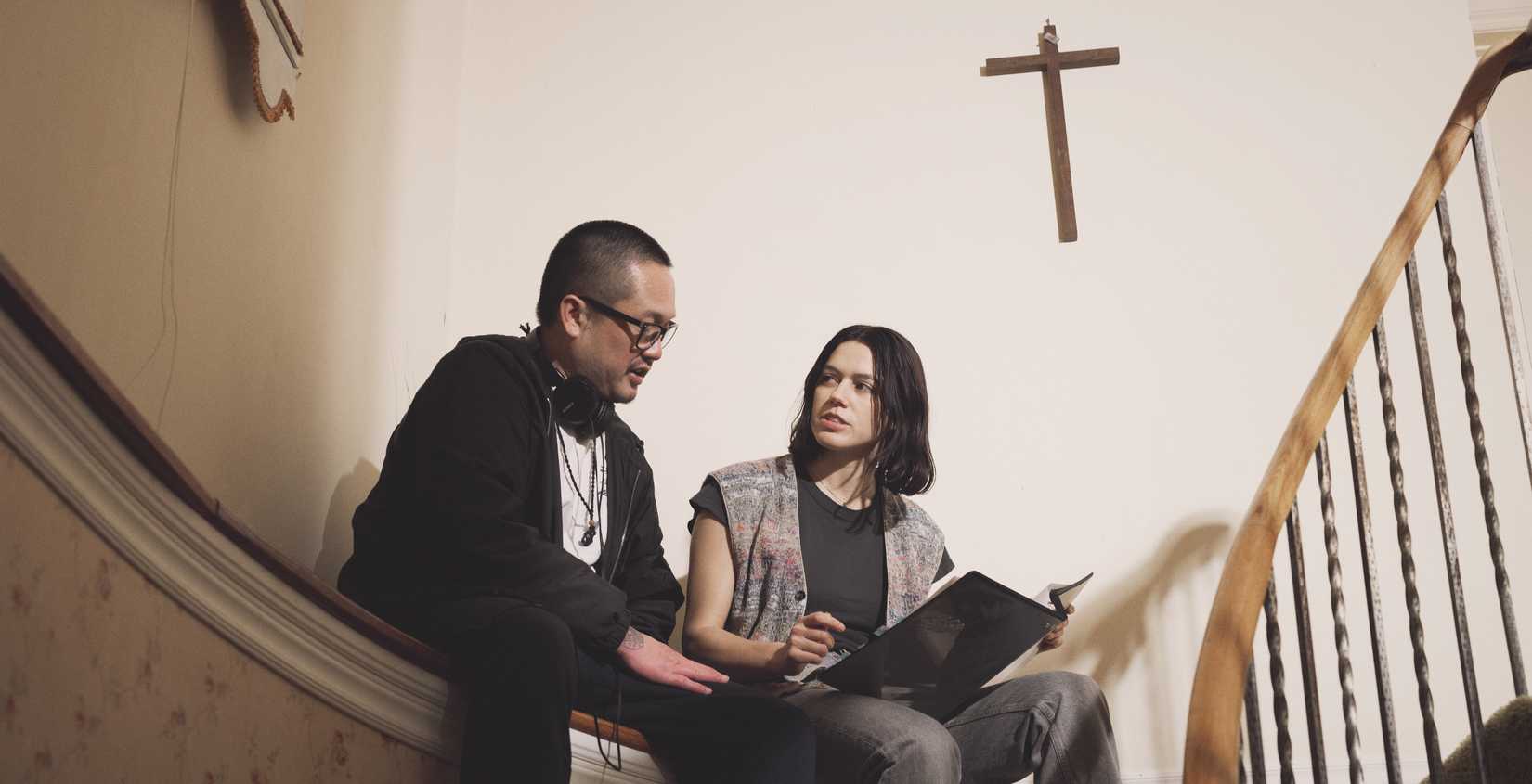 Director Ian Tuason and star Nina Kiri sit on a shelf above a curved stairway and consult a script on the set of Undertone