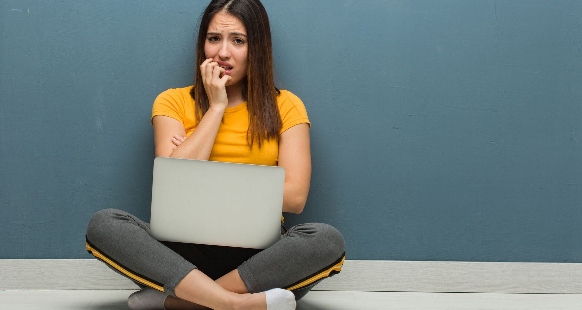 Young woman sitting on the floor with a Windows laptop biting nails, nervous and very anxious
