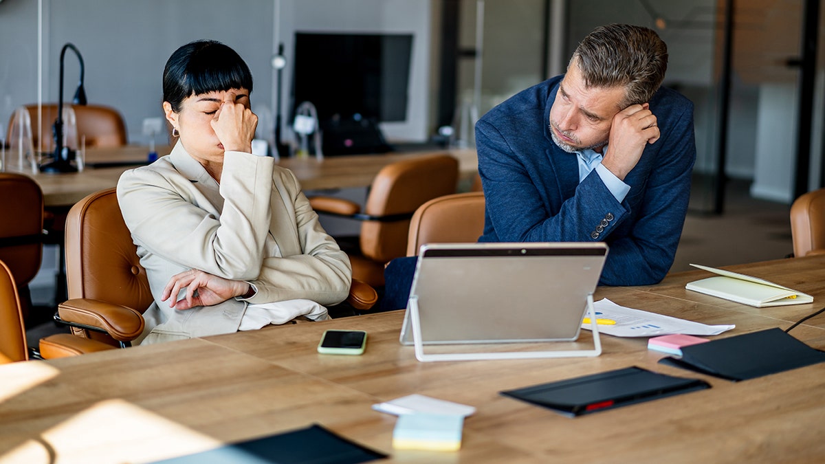 Two business professionals looking stressed and fatigued while working diligently at their office desks, surrounded by paperwork and devices