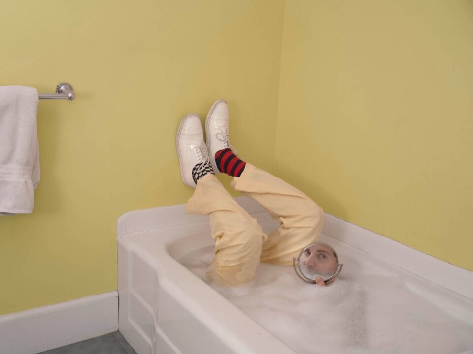 a surreal portrait by photographer Ben Zankn depicting a figure fully clothed in a bathtub with bubbles, looking into a mirror
