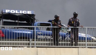 Two police officers leaning over a white railing outdoors. Behind them is a blue police vehicle and a police tent