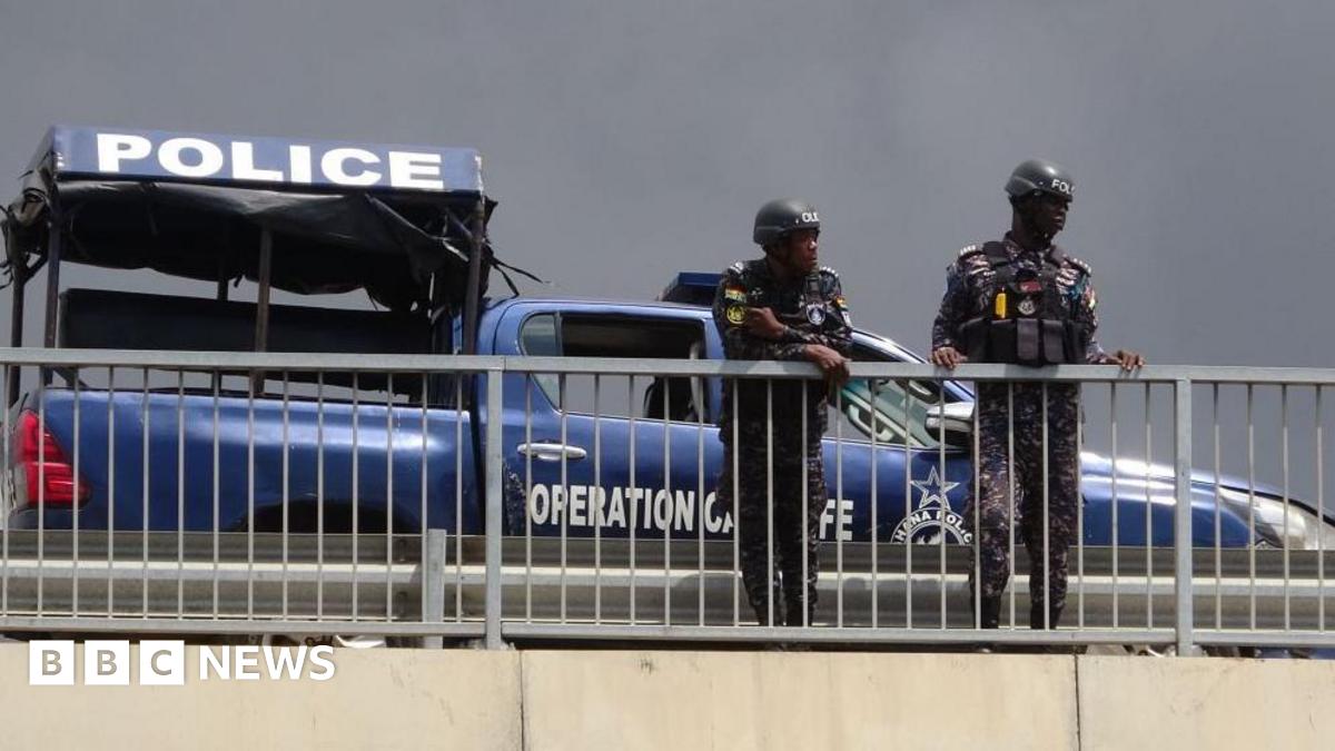 Two police officers leaning over a white railing outdoors. Behind them is a blue police vehicle and a police tent