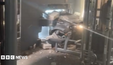 The front of a silver or grey car can be seen coming through a hole in the wall of the gym. Bricks and debris are lying around. Some gym equipment can be seen, including one apparatus which is right by where the car ended up.