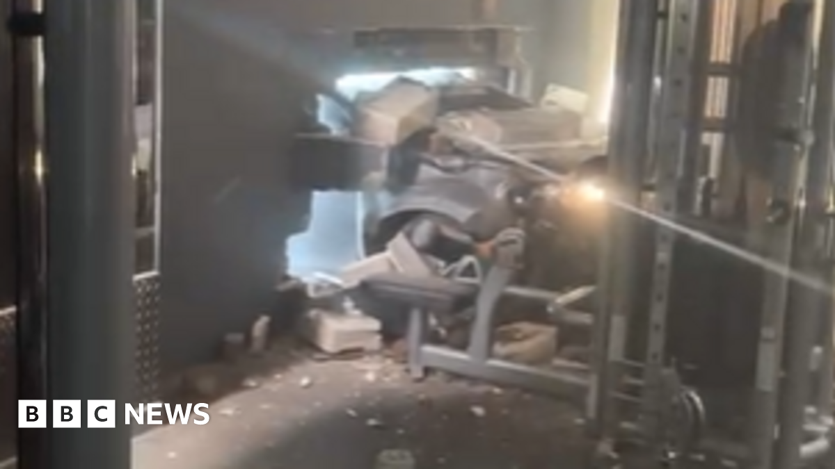 The front of a silver or grey car can be seen coming through a hole in the wall of the gym. Bricks and debris are lying around. Some gym equipment can be seen, including one apparatus which is right by where the car ended up.