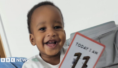 Noah Sibanda has short curly black hair and is smiling. He has a white jumper on and is holding a card that says "today I am 11 months old"