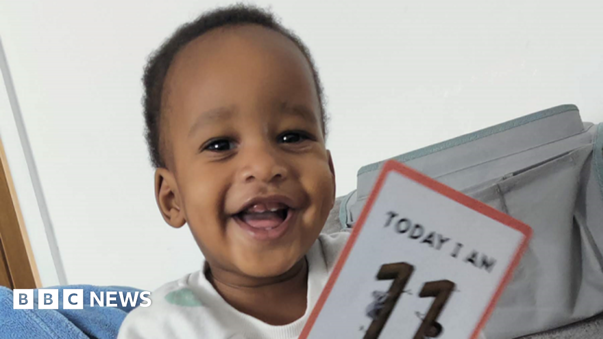 Noah Sibanda has short curly black hair and is smiling. He has a white jumper on and is holding a card that says "today I am 11 months old"